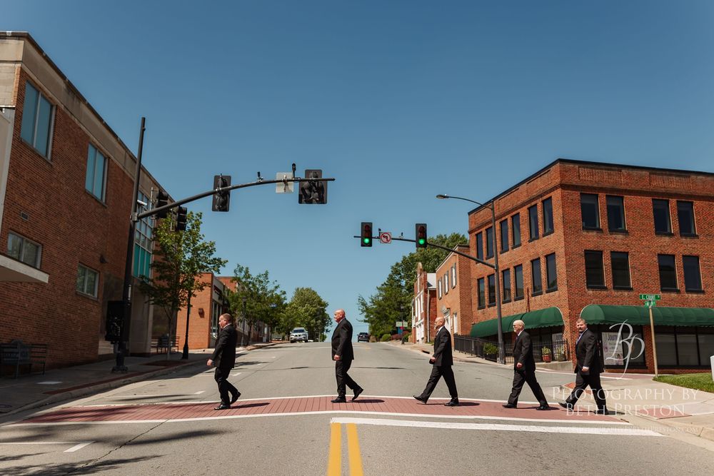 Pedestrians cross an intersection with traffic lights and brick buildings on a sunny day in a downtown area.