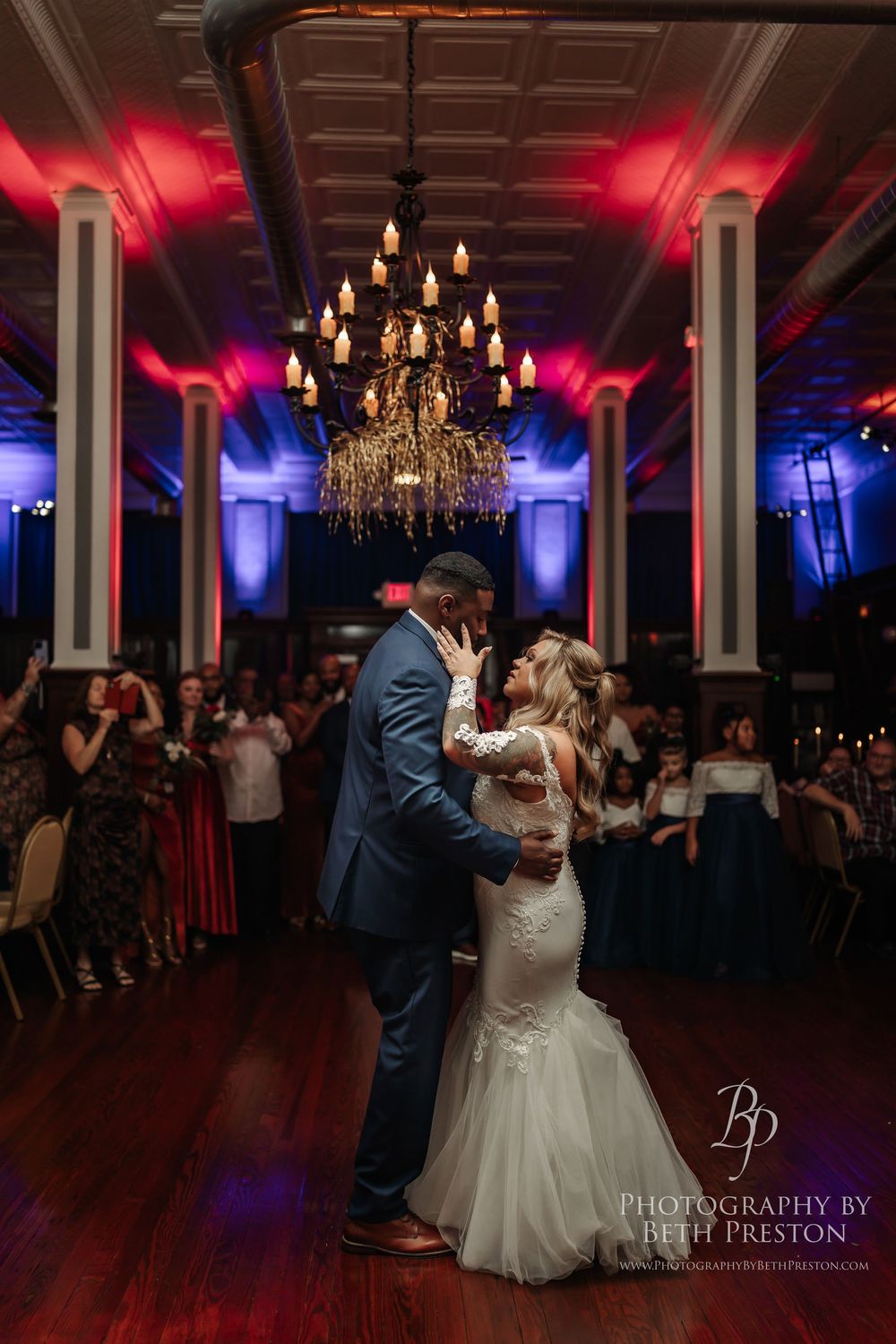 A couple shares their first dance under an elegant crystal chandelier with dramatic red and blue lighting at their wedding reception.