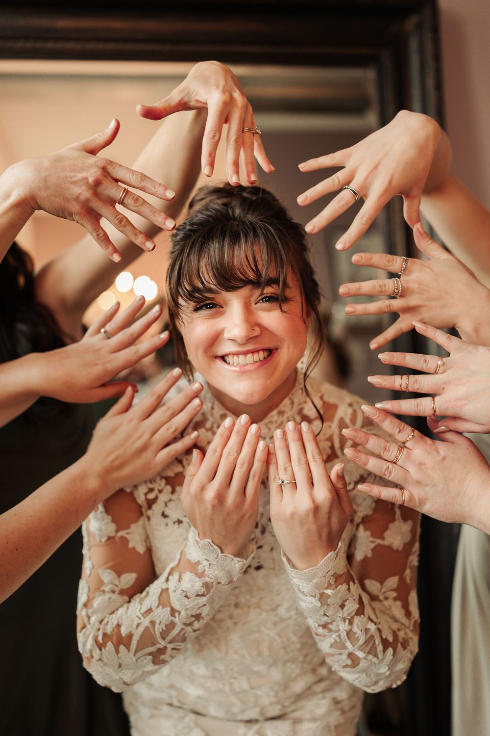 A series of hands showing off wedding rings while a bride in a lace dress smiles in the background.