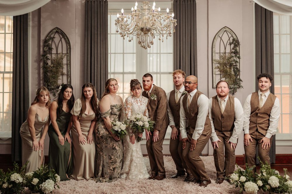 Wedding party poses in elegant venue with chandelier and arched windows.