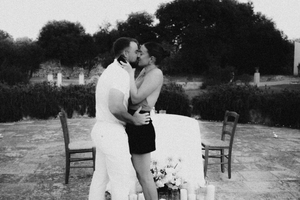Black and white photo of a romantic couple sharing a kiss at an outdoor table with chairs.