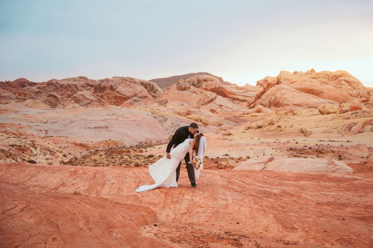 A couple in wedding attire embraces against a backdrop of red rock desert formations at sunset.