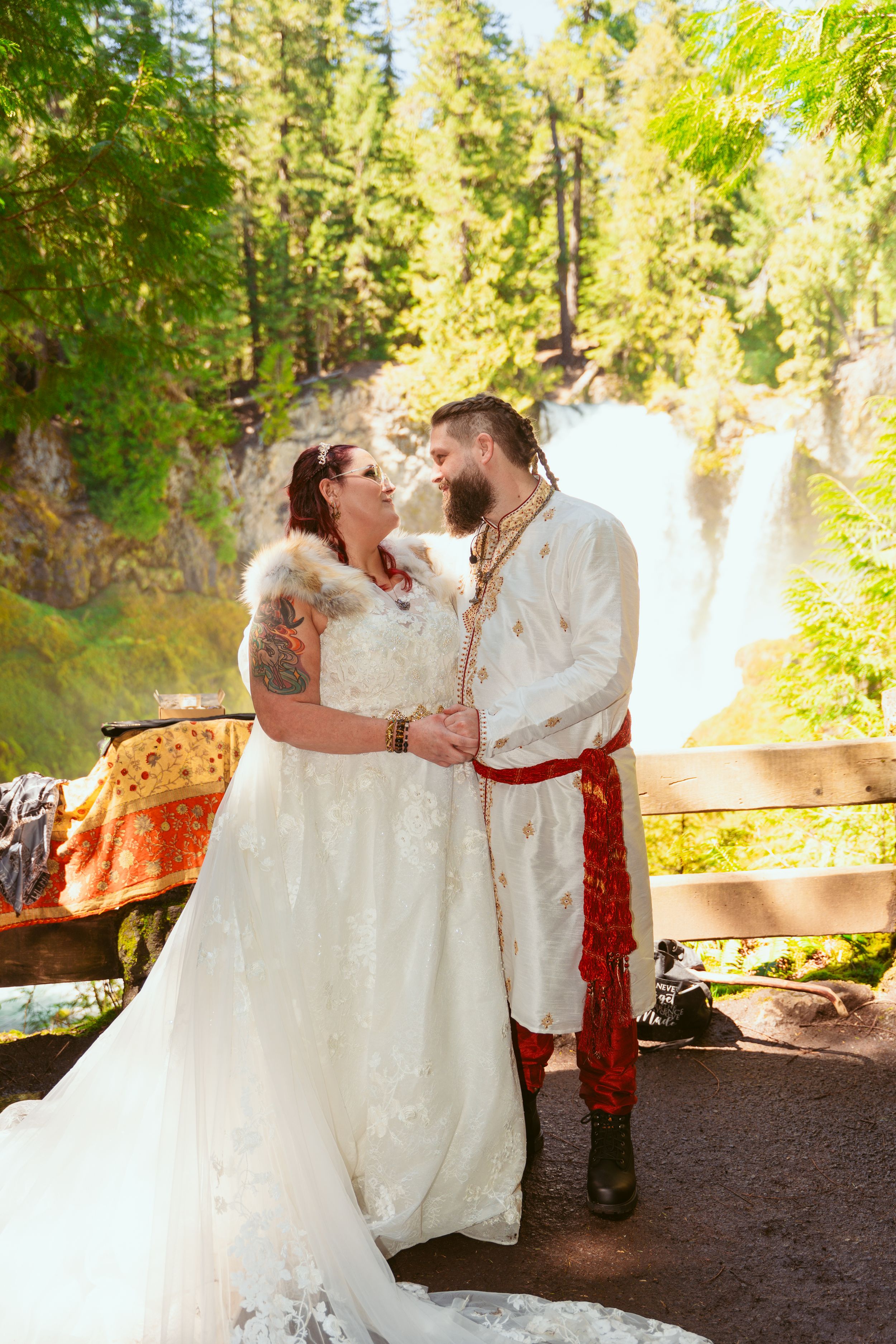 Newlyweds in white and red medieval costumes share intimate moments in a sunlit forest.