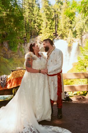 Romantic wedding portrait with majestic waterfall backdrop and traditional white and red ceremonial attire.