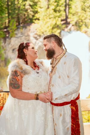 A romantic wedding couple wearing white attire share an intimate moment in a sunlit forest setting.