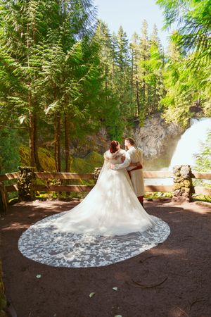 A romantic wedding scene with flowing white dress train against evergreen forest and waterfall backdrop.