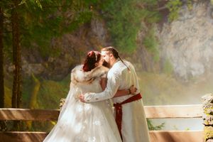 A romantic couple in elegant wedding attire share an intimate moment on a rustic wooden bridge surrounded by lush greenery.