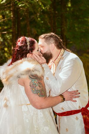 A romantic wedding couple in medieval inspired attire sharing a tender kiss outdoors with a green forest backdrop.