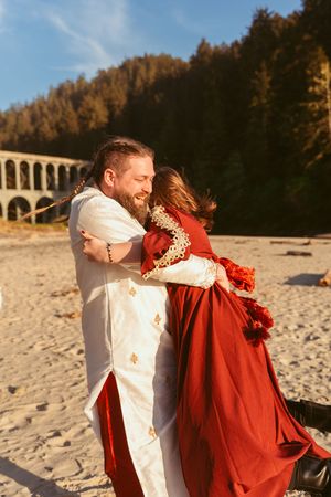 A couple in formal attire dances joyfully on a beach with a viaduct and forested hills in the background.