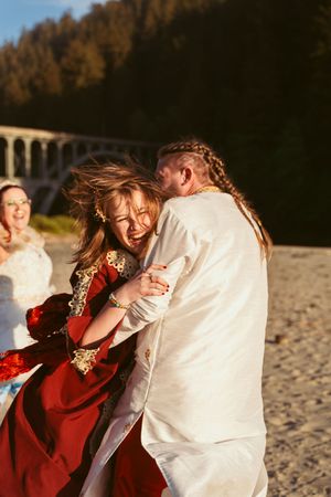 A playful beach wedding scene with a couple in white and red attire dancing together near an arched bridge.