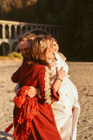Dancing couple laughs joyfully on beach with sunset and arched bridge in background.