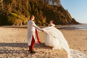 A couple in wedding attire dances playfully on a sandy beach with cliffs and ocean views.