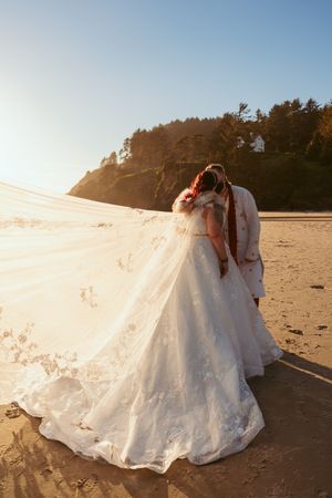 Romantic couple embraces on a sunlit beach with lush forest background and flowing white dress in golden hour light.