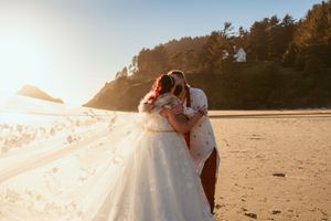 Two people embrace on a sandy beach at sunset with sunlight streaming through a flowing white dress.