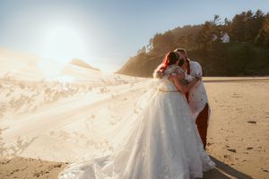 A bride's long white veil flows elegantly in the wind along a scenic beach at sunset.