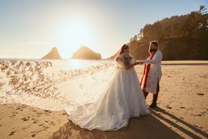 A couple in wedding attire pose against dramatic coastal rock formations at sunset on a beach.