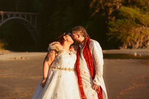 A couple in wedding attire shares a romantic moment on a beach at sunset with a bridge in the background.