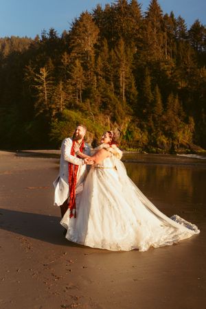 Newlyweds dancing joyfully on a beach with tall evergreen trees in the background.