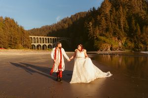Sunset wedding photo on beach with historic bridge in background showing full length traditional attire.