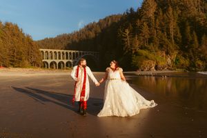 Couple holds hands on a sunset-lit beach with Cape Creek Bridge in background at Heceta Head Beach, Oregon coast.
