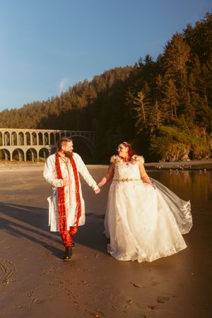 A couple in wedding attire walk hand in hand on a beach at sunset with a stone bridge visible in the background.