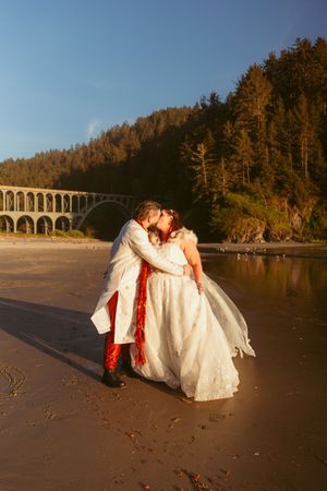 A romantic beach wedding scene with an arched bridge and coastal forest backdrop.