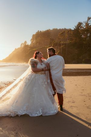 Newlyweds share intimate moment on beach at golden hour with forest cliffs in background.