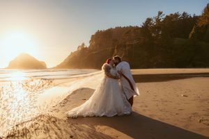 Couple embraces on scenic beach at sunset with rocky coastline and glowing sun over Pacific Ocean waters.