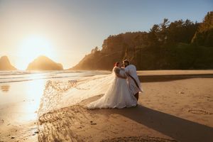 A couple in wedding attire embraces on a scenic beach at sunset with towering cliffs and rocky sea stacks in the background.