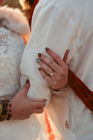 Close-up of hands showing engagement ring and jewelry details against white wedding attire and orange sunset lighting.