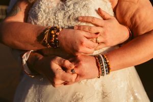 Close-up of hands with wooden and beaded bracelets embracing against white lace wedding dress fabric.