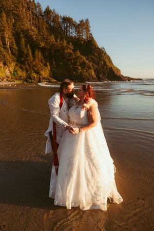 A couple in wedding attire walks together on a scenic beach with cliffs and evergreen trees in the background.