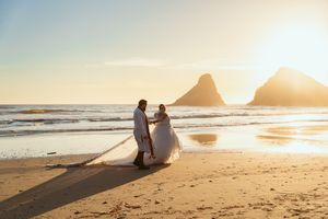 Silhouetted newlyweds standing on a beach at sunset with dramatic mountain peaks in the background.