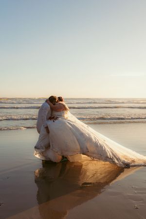 A romantic beach wedding photo with newlyweds embracing at sunset as waves reflect their moment in the wet sand.