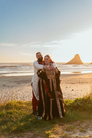A couple embraces on a scenic beach at sunset with rock formations and ocean waves in the background.