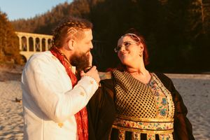 A couple in vintage-inspired wedding attire sharing a playful moment on a beach with a bridge in the background.