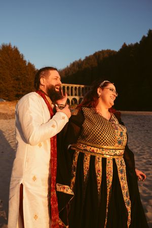 Wedding party in white and patterned medieval costumes posing playfully at sunset on a beach.