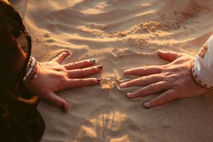 Two hands reaching towards each other on sandy beach at golden hour with wedding rings visible.