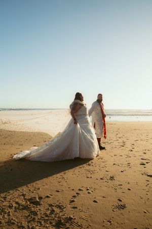 A couple in wedding attire walking on a sandy beach during sunset with ocean waves in the background.