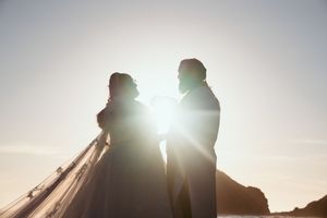 Dramatic backlit silhouettes of wedding couple embracing on beach during golden hour sunset.
