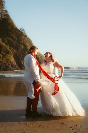 A romantic couple in wedding attire sits together playfully on a sunny beach with cliffs in the background.
