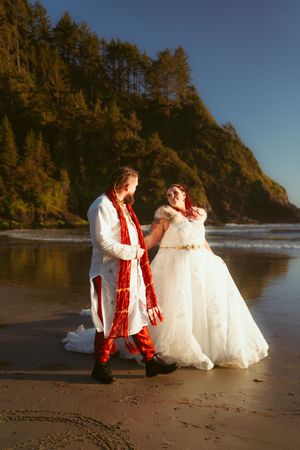 A couple shares a romantic moment on a sunset-lit beach with dramatic cliffs and evergreen trees in the background.