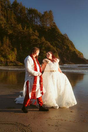 Two figures in white and red traditional attire pose romantically on a scenic beach at sunset with cliffs in background.