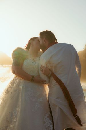 A romantic wedding couple shares a kiss at sunset, silhouetted against the golden light in elegant wedding attire.