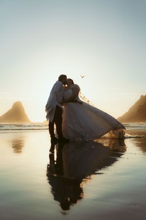 Silhouetted couple in wedding attire embrace on reflective beach waters during golden hour with rocky coastline behind them.