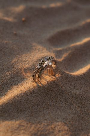 Diamond engagement ring nestled in golden sand with warm sunlight creating shadows and texture.