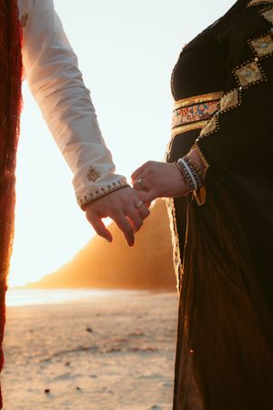 A couple holds hands against a glowing sunset background on a beach, wearing traditional ethnic clothing.