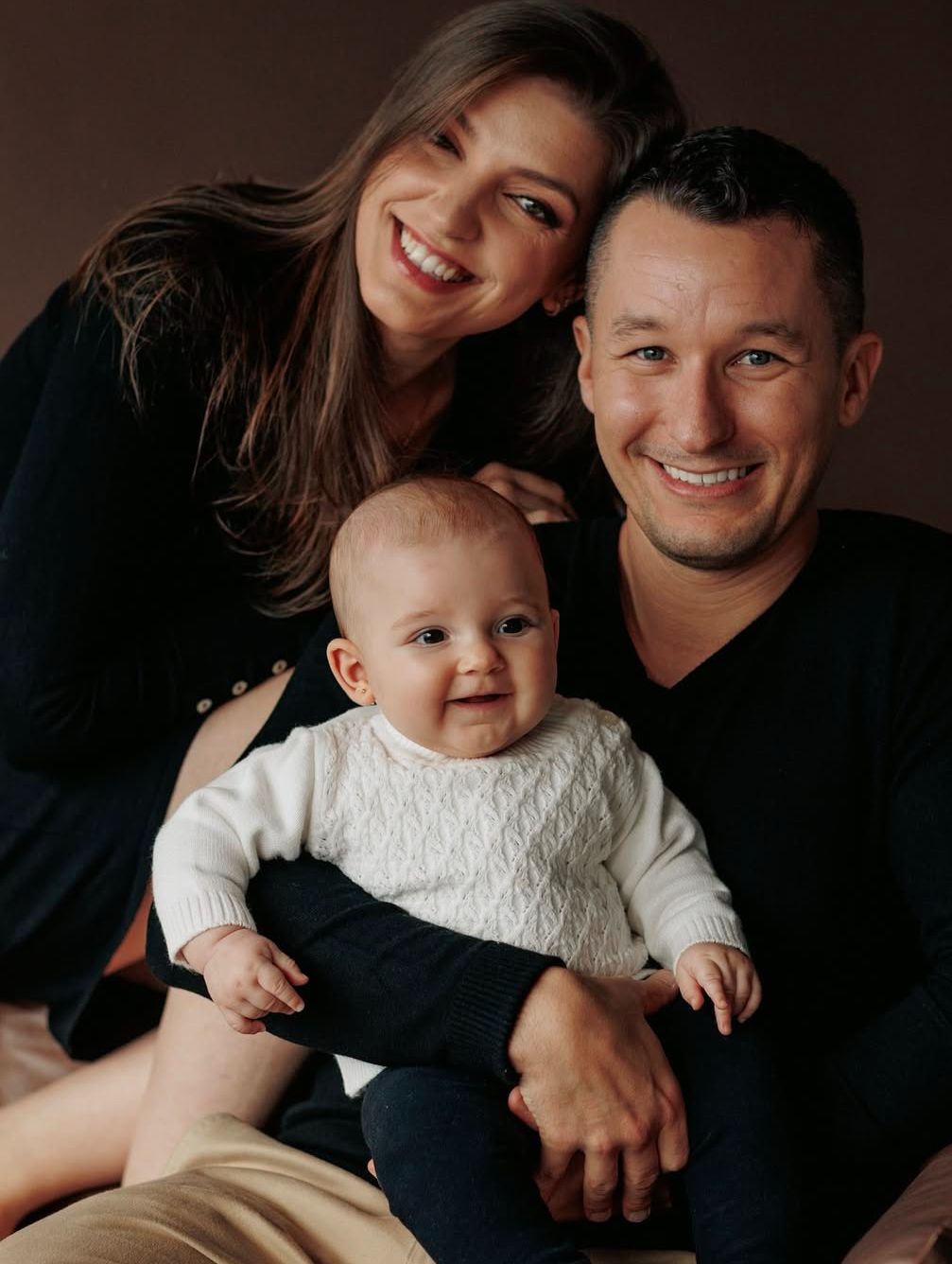 Studio portrait of family with baby wearing white sweater against dark background.