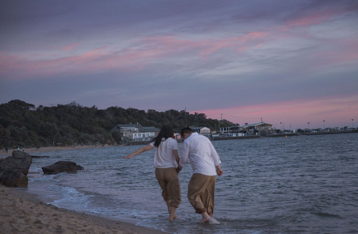 A couple embraces on a coastal cliff with ocean waves and golden grass during sunset.