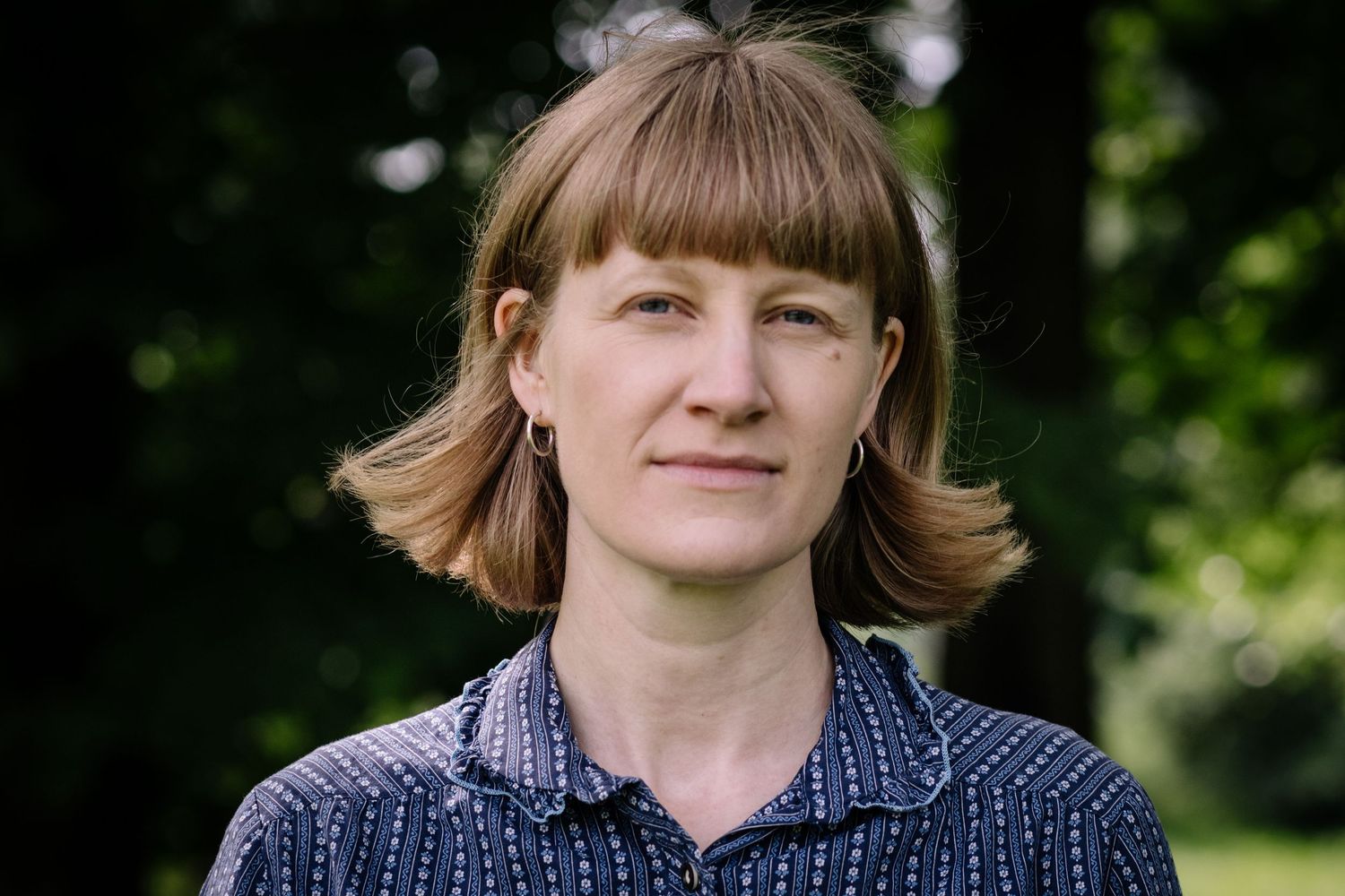 Close-up outdoor portrait of someone with flipped shoulder-length hair and bangs wearing a patterned navy blue shirt.